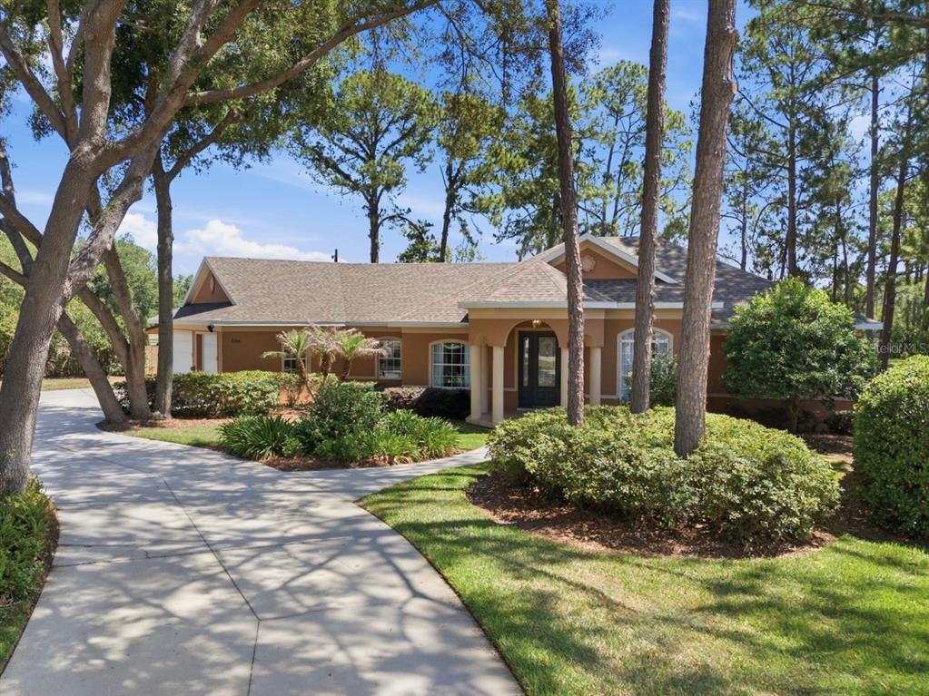 A single-story house with a tan stucco exterior and a concrete driveway sits among trees — 1064 CEASARS COURT, MOUNT DORA, FL