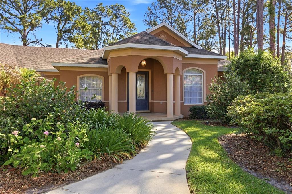 A concrete walkway leads to the arched, columned entrance of a tan stucco house — 1064 CEASARS COURT, MOUNT DORA, FL