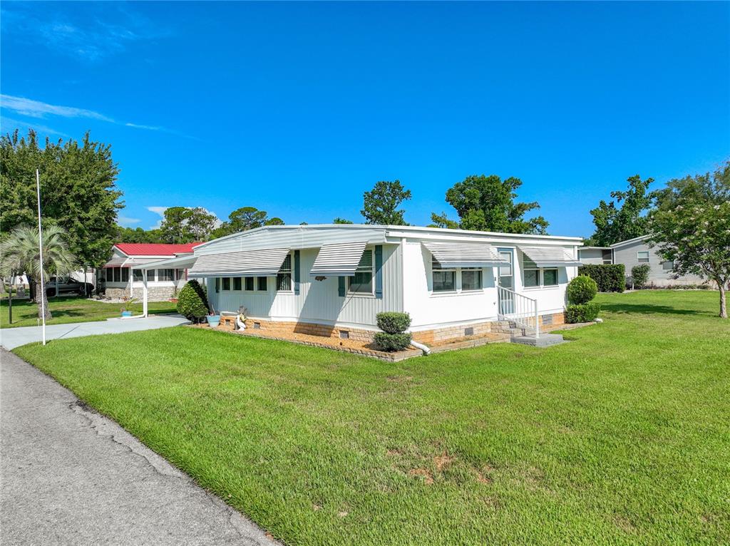 A white mobile home with metal awnings sits on a grassy lot under a blue sky — 1925 BROOKSIDE DRIVE, MOUNT DORA, FL