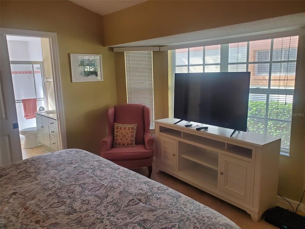A bedroom features a pink armchair, a white media console with a television, and an ensuite — 1232 CAPRI ISLES BOULEVARD UNIT 48, VENICE, FL
