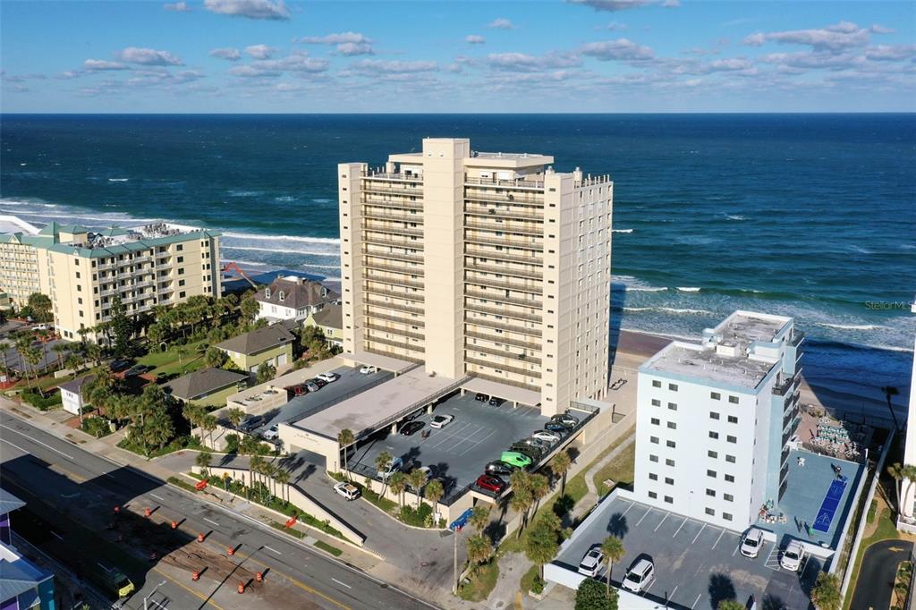 An aerial view shows a tall beachfront condominium building with an attached parking lot — 89 S ATLANTIC AVENUE UNIT 1002, ORMOND BEACH, FL