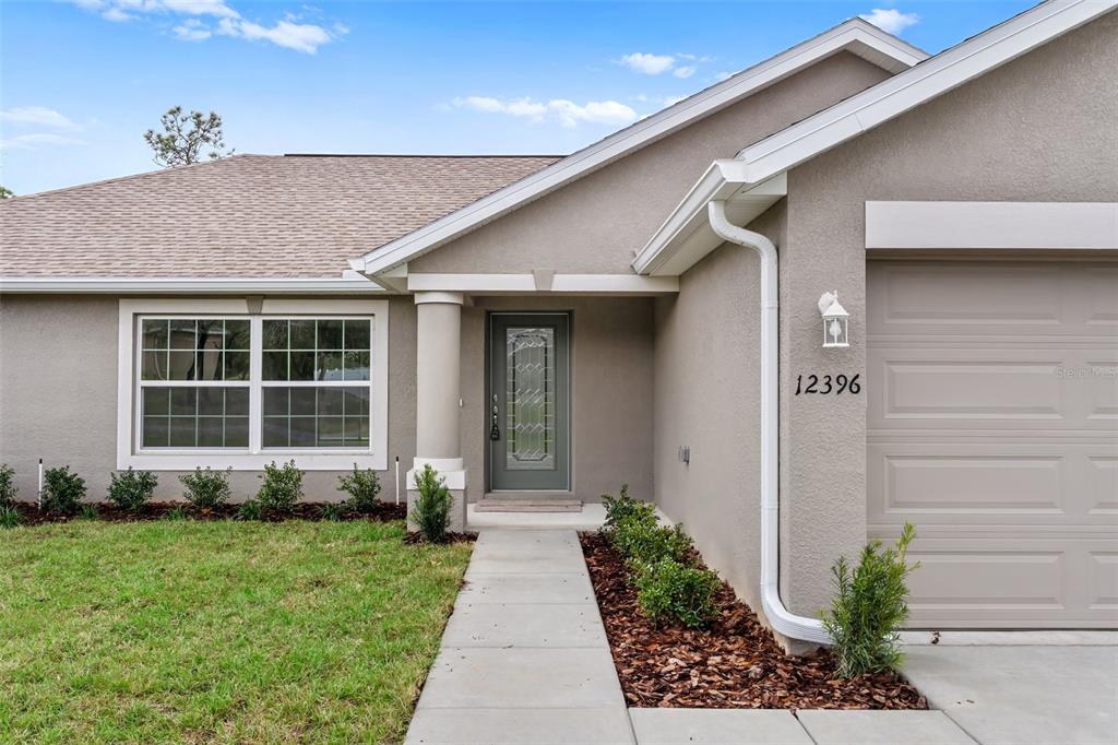 A single-story house exterior features a gray stucco facade, a front door, and a garage — 12073 MANASSAS AVENUE, WEEKI WACHEE, FL