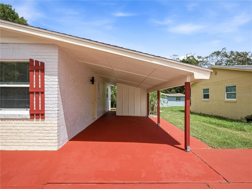 A covered carport with a red concrete floor sits beside a white brick house — 629 CEDAR PARK DRIVE, DAYTONA BEACH, FL