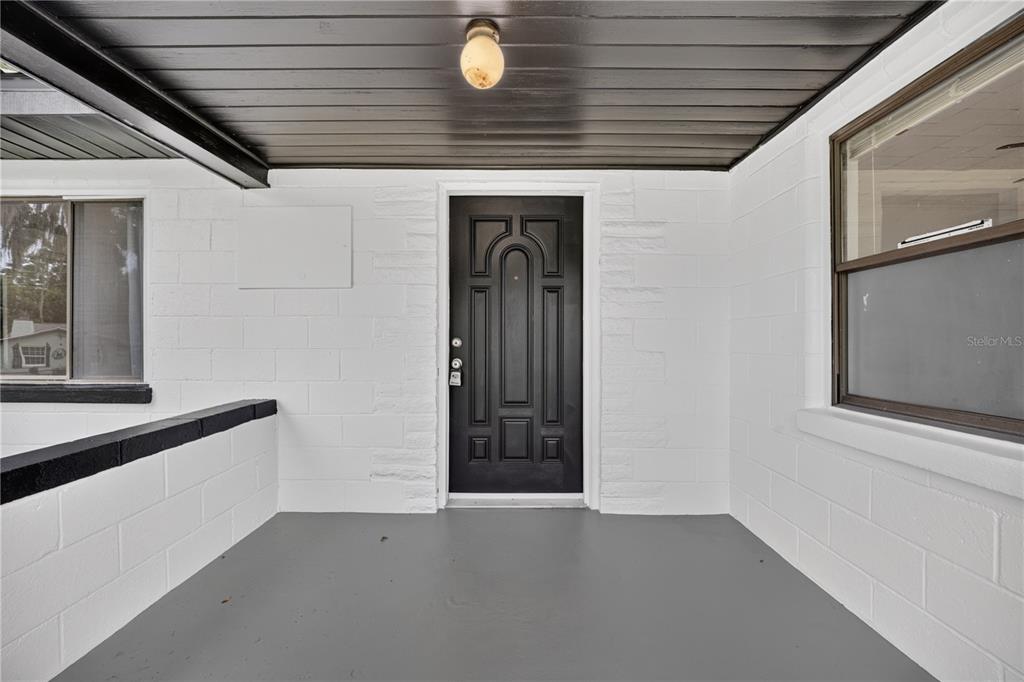 A covered porch with a black front door, white block walls, and gray painted flooring — 5514 SE HAMES ROAD, BELLEVIEW, FL