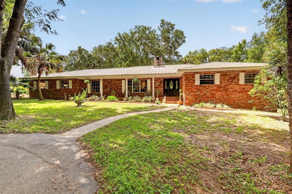 A single-story brick house with a metal roof sits on a grassy lot with trees — 22740 COUNTY ROAD 44A, EUSTIS, FL