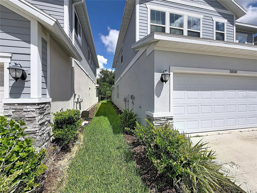 A narrow grassy side yard sits between two gray two-story houses with white garage doors — 28591 COZY CREEK DRIVE, WESLEY CHAPEL, FL