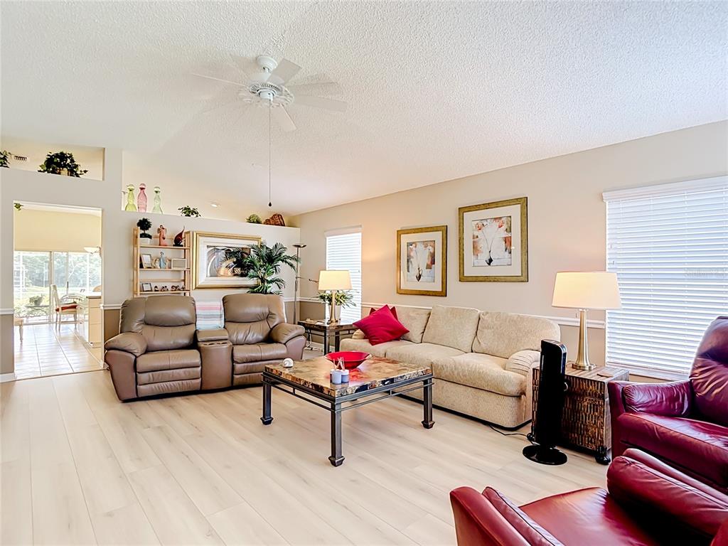 A living room features light wood-look flooring, a ceiling fan, and various seating arrangements — 7828 CRAIGHURST LOOP, NEW PORT RICHEY, FL