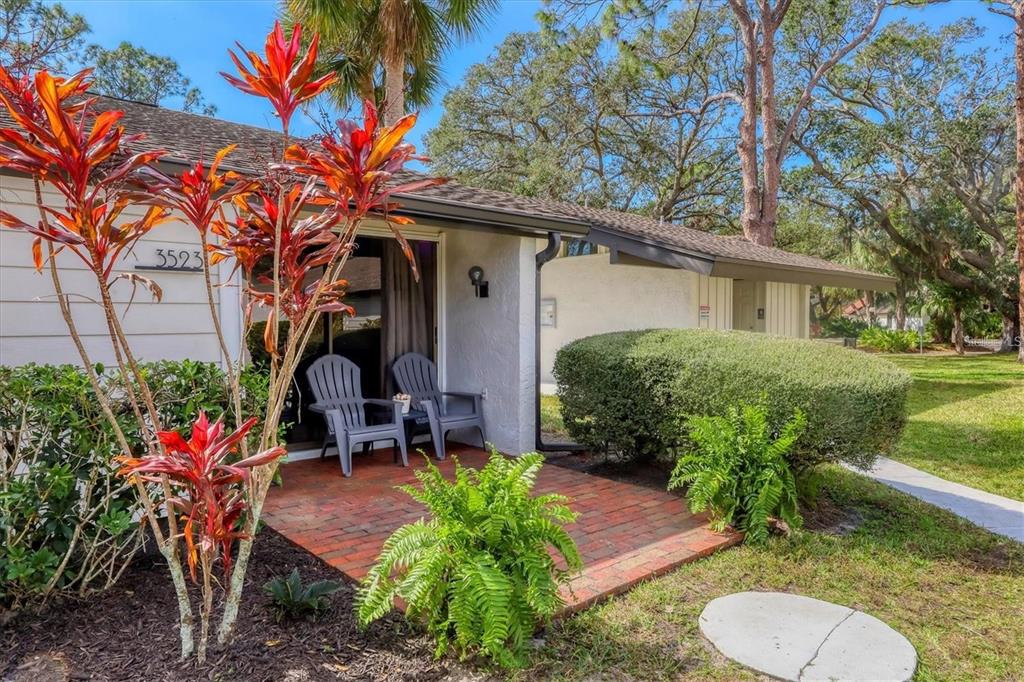 A brick patio with two chairs sits in front of a house with red-leafed plants — 3523 LONGMEADOW UNIT 12, SARASOTA, FL