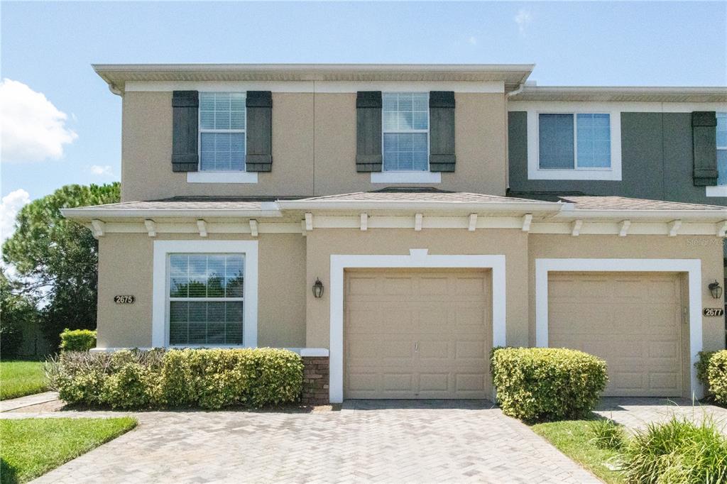 A two-story townhouse exterior with a tan stucco facade, two garage doors, and landscaped bushes — 2675 RIVER LANDING DRIVE, SANFORD, FL