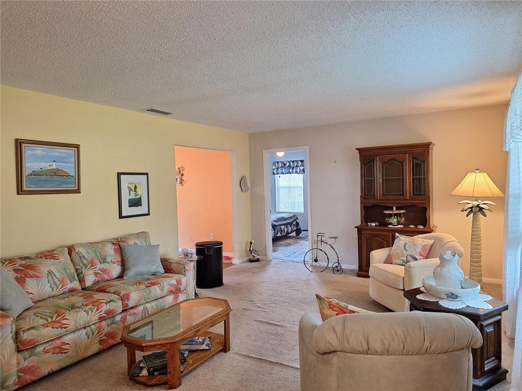 A living room features a floral sofa, beige armchairs, a wooden hutch, and carpeted floors — 2707 PINE TREE DRIVE, EDGEWATER, FL