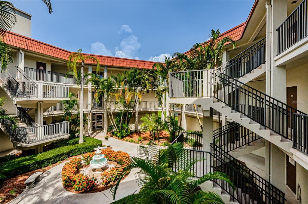 An outdoor courtyard with a central fountain, palm trees, and multi-level apartment building walkways — 2700 BAYSHORE BOULEVARD UNIT 2201, DUNEDIN, FL