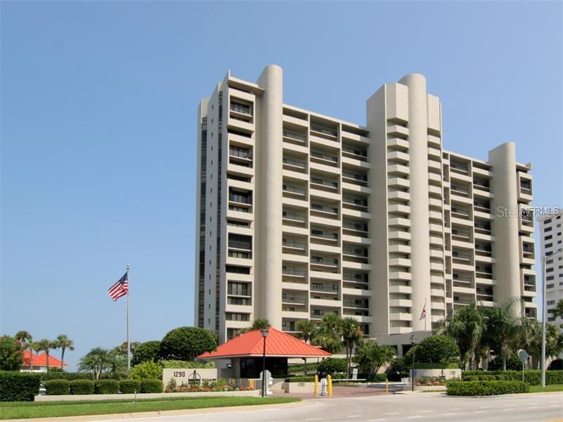 A tall, beige multi-story residential building with a gated entrance and a flag on site — 1290 GULF BOULEVARD UNIT 1003, CLEARWATER BEACH, FL