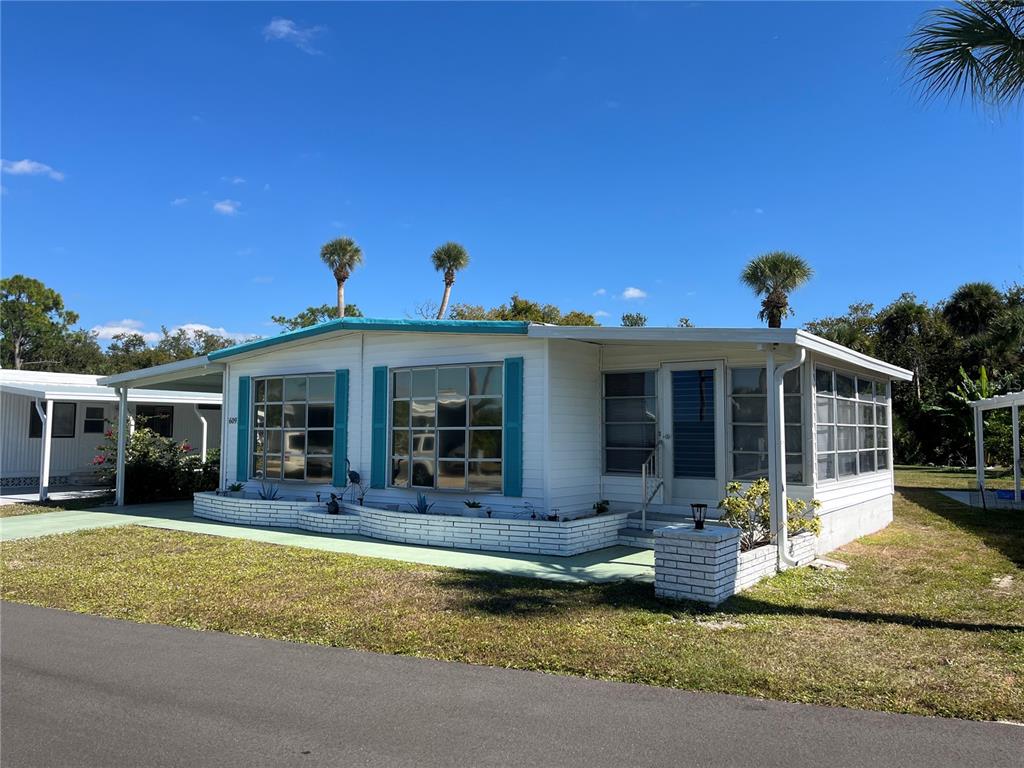 A white manufactured home with teal shutters and a brick planter sits on a lawn — 609 CERVINA DRIVE, VENICE, FL