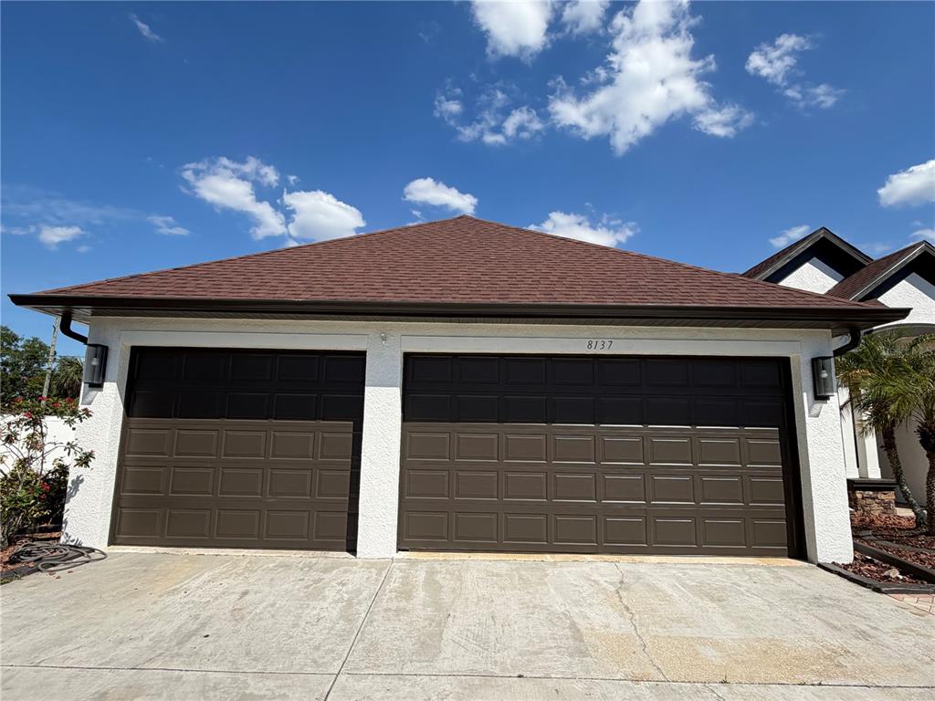 A two-car garage with brown paneled doors and a hip roof under a blue sky — 8137 GOLDEN BEAR LOOP, PORT RICHEY, FL