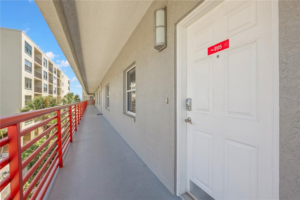 An outdoor apartment walkway features a white door labeled 205 and a red metal railing — 12935 GULF LANE UNIT 205, MADEIRA BEACH, FL
