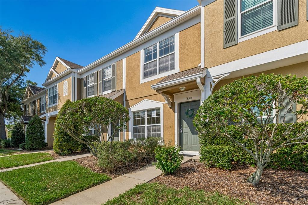A two-story townhouse exterior with tan stucco walls, a green front door, and landscaping — 1330 STANDRIDGE DRIVE, WESLEY CHAPEL, FL