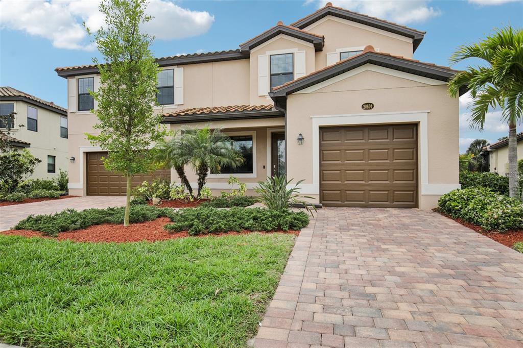A two-story suburban house with a tan stucco exterior, two garage doors, and paver driveway — 20604 BENISSIMO DRIVE, VENICE, FL