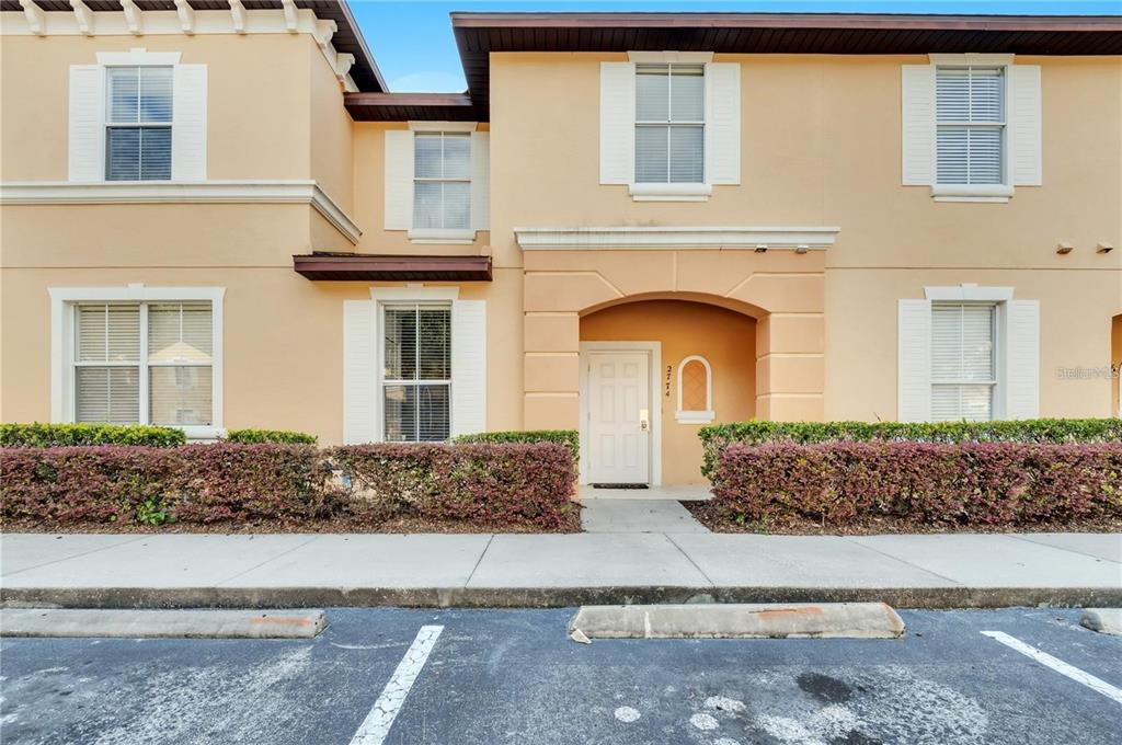 A two-story tan townhouse exterior features a white front door and a paved parking area — 2774 CORVETTE LANE, KISSIMMEE, FL