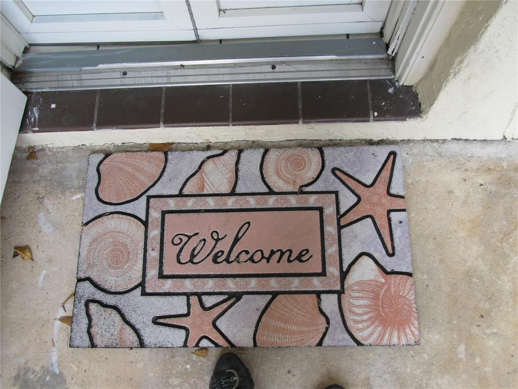 A coastal-themed welcome mat sits on a concrete porch in front of a glass door — 7050 W COUNTRY CLUB DRIVE N UNIT 10-A, SARASOTA, FL