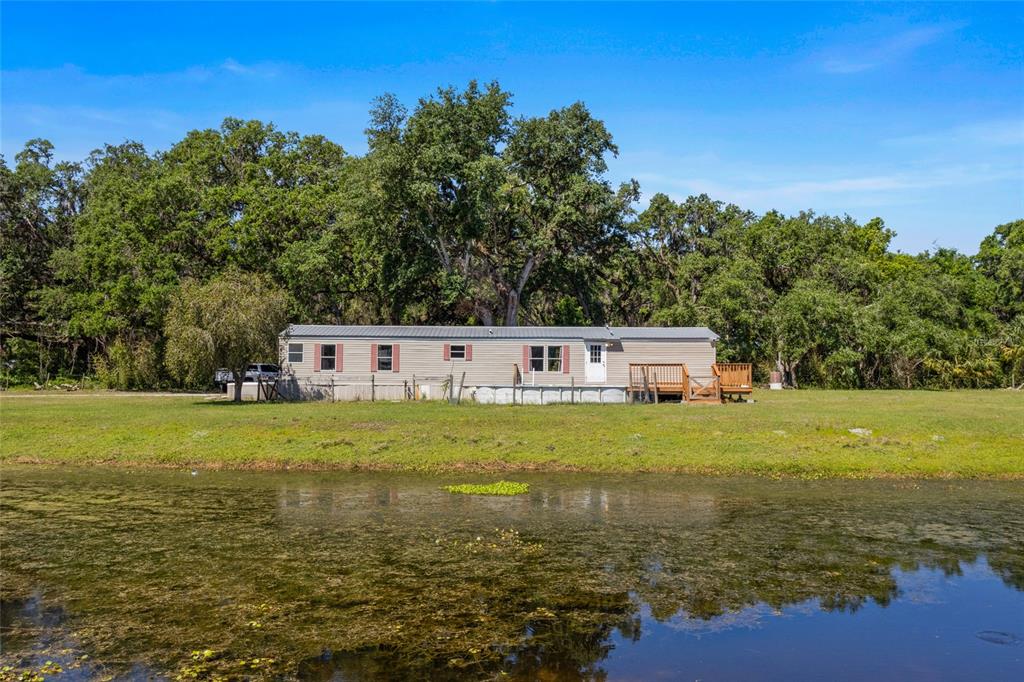 A single-story manufactured home sits on a grassy lot beside a pond with trees behind — 11902 CR 727, WEBSTER, FL