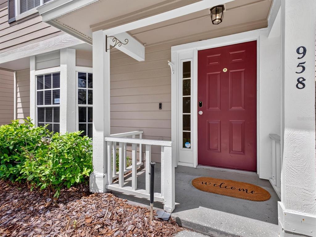 A red front door with a welcome mat sits under a covered porch with white railings — 958 SYKES COURT UNIT 104, ORLANDO, FL