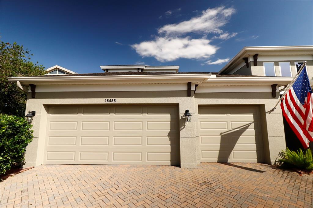 A beige two-car garage with a brick paver driveway and an American flag — 16485 COURTYARD LOOP, LAND O LAKES, FL