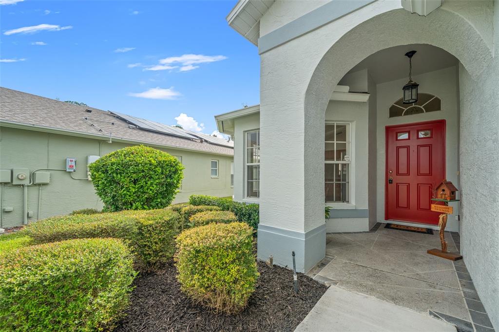 A red front door is set within a covered entryway next to manicured green shrubs — 18208 BRELAND DRIVE, HUDSON, FL