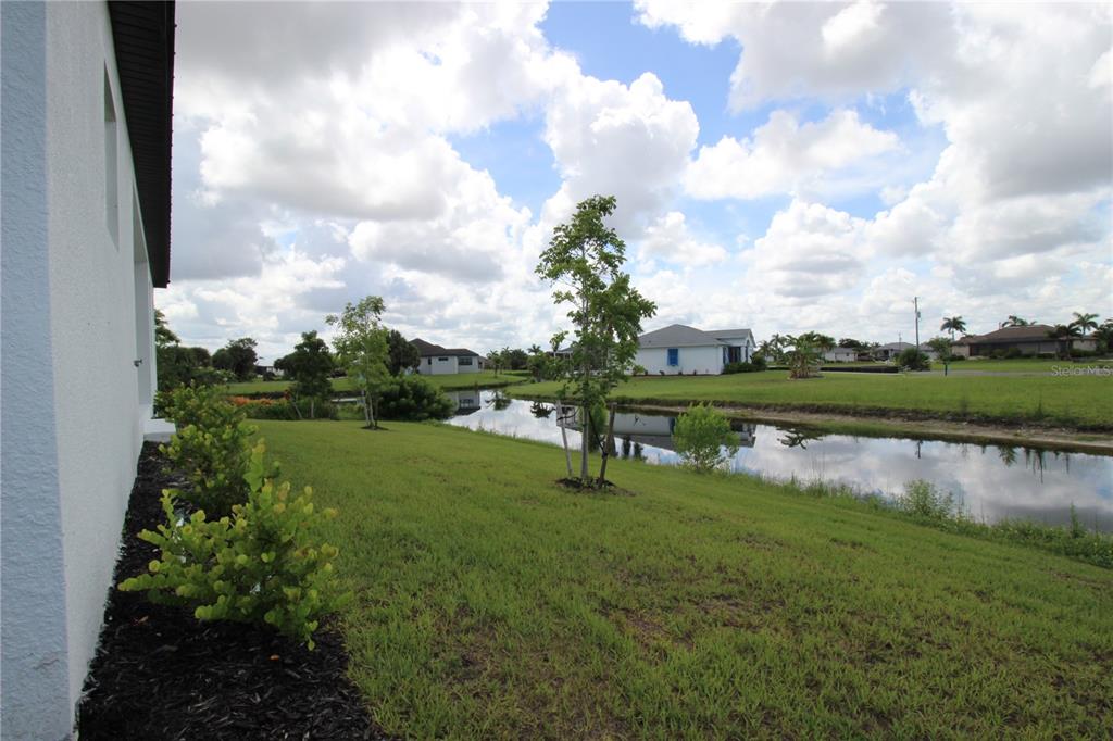 A grassy backyard slopes down to a canal with several young trees and distant houses — 17030 ESPANA CIRCLE, PUNTA GORDA, FL