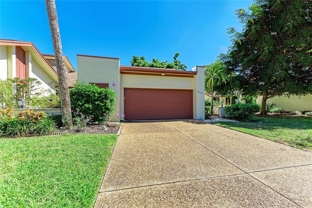 A single-story house exterior features a two-car garage, a concrete driveway, and surrounding landscaping — 6207 COURTSIDE DRIVE UNIT 23, BRADENTON, FL