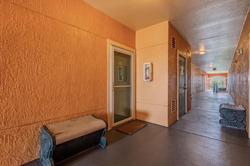 An outdoor apartment hallway with orange stucco walls, a front door, and a small bench — 2757 VIA CIPRIANI UNIT 1131B, CLEARWATER, FL
