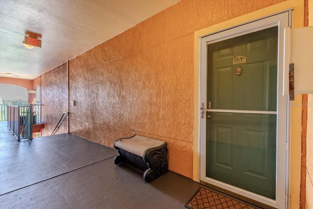 An outdoor apartment walkway features a green door, a small bench, and an exit sign — 2757 VIA CIPRIANI UNIT 1131B, CLEARWATER, FL
