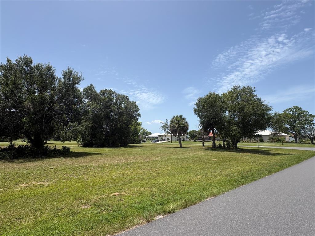 A grassy vacant lot with several mature trees sits next to a paved road — 16842 ACAPULCO ROAD, PUNTA GORDA, FL