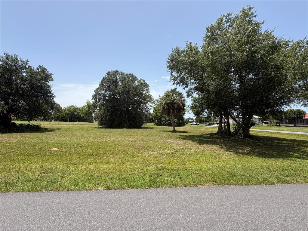 A grassy, vacant lot features several large trees under a clear blue sky — 16842 ACAPULCO ROAD, PUNTA GORDA, FL