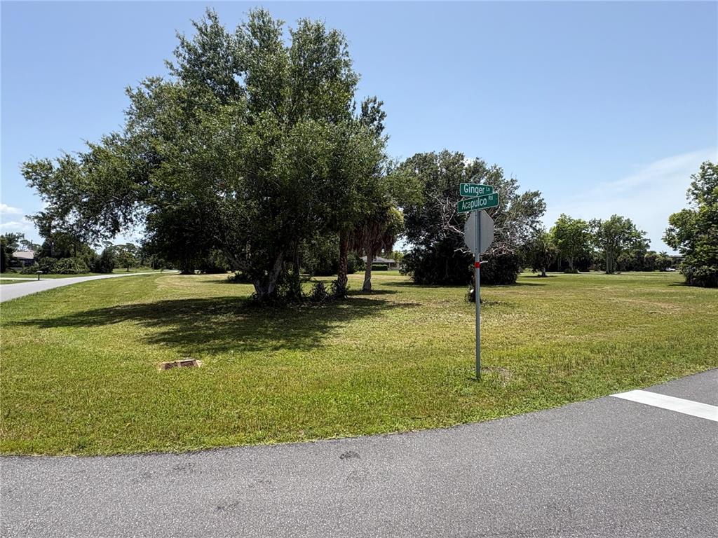 A street sign stands at the corner of a grassy lot with several large trees — 16842 ACAPULCO ROAD, PUNTA GORDA, FL