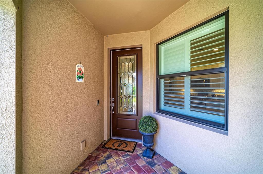 A covered entryway features a brown front door with decorative glass, a window, and pavers — 17648 NORTHWOOD PLACE, LAKEWOOD RANCH, FL