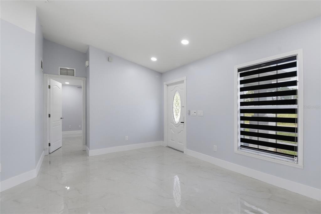 An empty room features light blue walls, white trim, marble-look tile flooring, and a front door — 1104 MORTON STREET E, LEHIGH ACRES, FL
