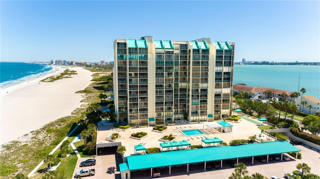 An aerial view shows a tall beachfront condominium building with a pool and parking structure — 1380 GULF BOULEVARD UNIT 805, CLEARWATER, FL
