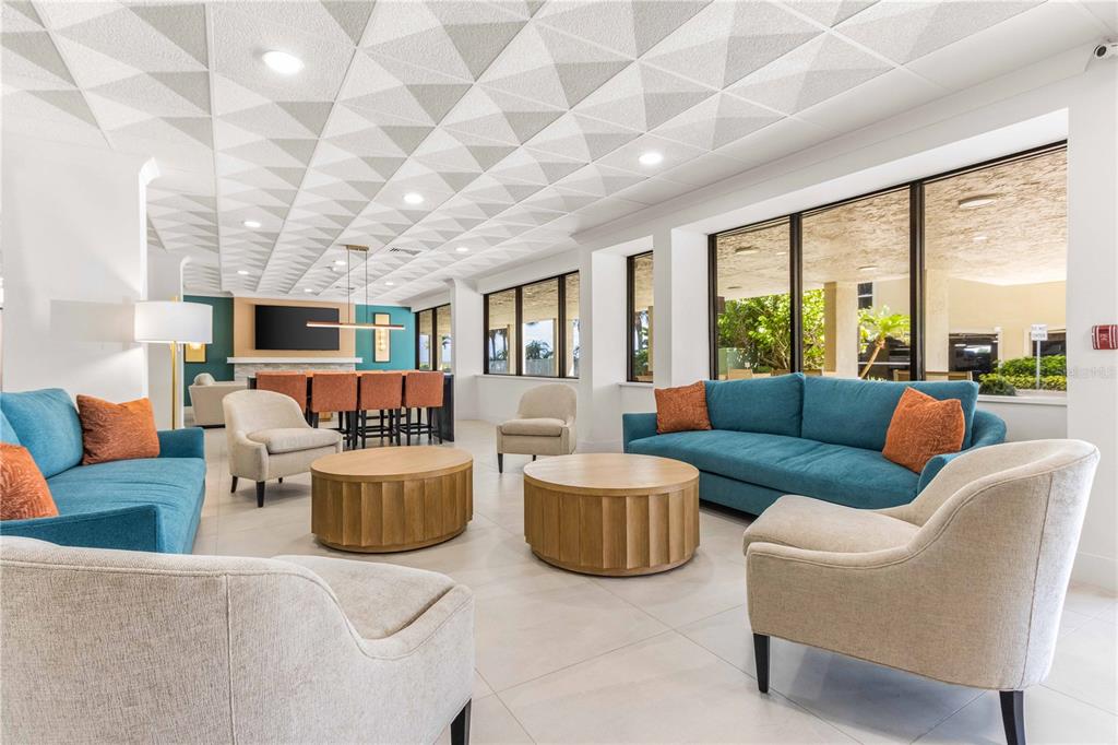 A modern lounge area features blue sofas, beige armchairs, round wooden tables, and geometric ceiling tiles — 1380 GULF BOULEVARD UNIT 805, CLEARWATER, FL