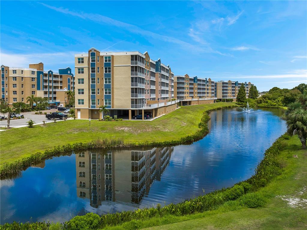 A multi-story apartment complex sits beside a pond with a fountain under a blue sky — 960 STARKEY ROAD UNIT 7205, LARGO, FL