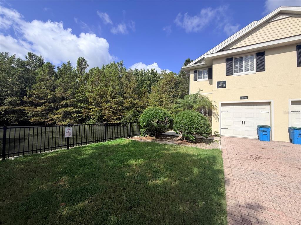 A two-story tan townhouse with a white garage door sits beside a grassy yard and pond — 10703 REGENT SQUARE DRIVE UNIT 1801, ORLANDO, FL