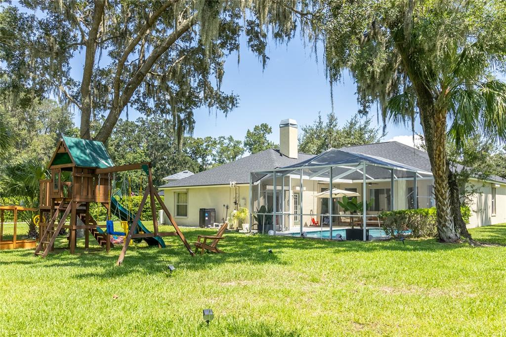 A backyard features a wooden playset, a screened-in pool, and large trees on a lawn — 12526 RIVER BIRCH DRIVE, RIVERVIEW, FL