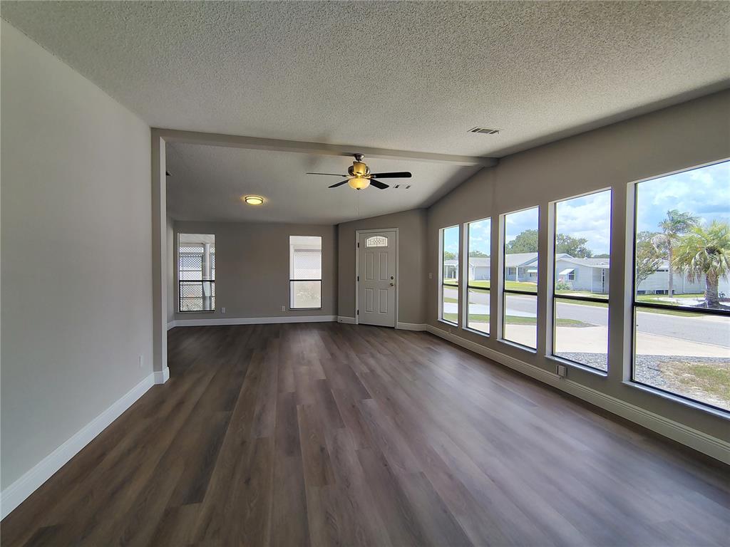 An empty living room features wood-look flooring, a ceiling fan, and large windows — 702 TURNBERRY LANE, LADY LAKE, FL
