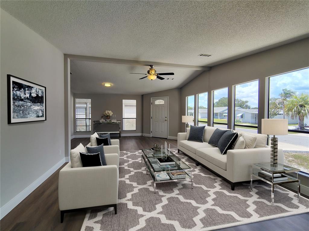 A living room features light-colored sofas, a patterned rug, and large windows overlooking a neighborhood — 702 TURNBERRY LANE, LADY LAKE, FL