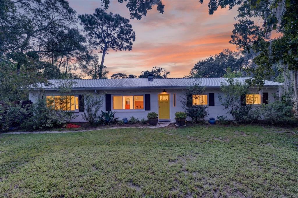 A single-story white house with a yellow front door sits behind a grassy lawn at sunset — 3929 NW 20TH TERRACE, GAINESVILLE, FL