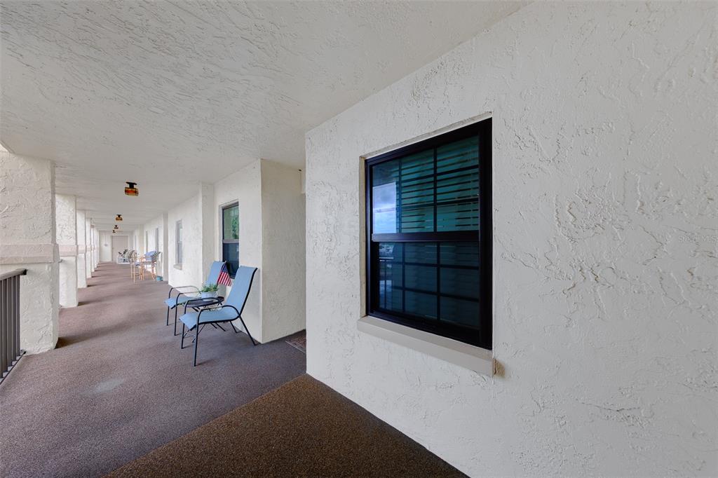 A covered outdoor walkway features textured white walls, a dark carpet, and blue patio chairs — 19029 US HIGHWAY 19 N UNIT 26-204, CLEARWATER, FL