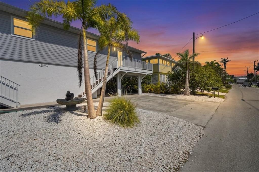 An elevated house with gray siding, palm trees, and a gravel yard at sunset — 3303 6TH AVENUE UNIT B, HOLMES BEACH, FL