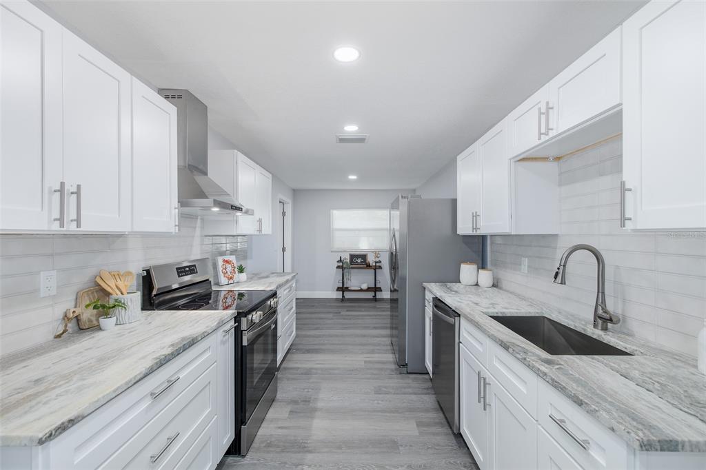 A galley kitchen features white cabinets, marble-patterned countertops, stainless steel appliances, and gray wood flooring — 1815 DRIFTWOOD CIRCLE S, OLDSMAR, FL