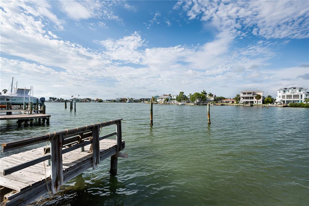 A wooden dock extends into a calm waterway with houses visible along the distant shoreline — 14035 E PARSLEY DRIVE, MADEIRA BEACH, FL