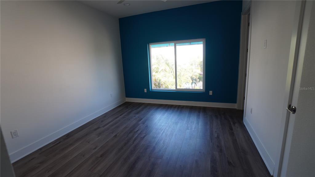 An empty bedroom features wood-look flooring, white walls, and a blue accent wall with a window — 15405 GULF BOULEVARD UNIT B2, MADEIRA BEACH, FL