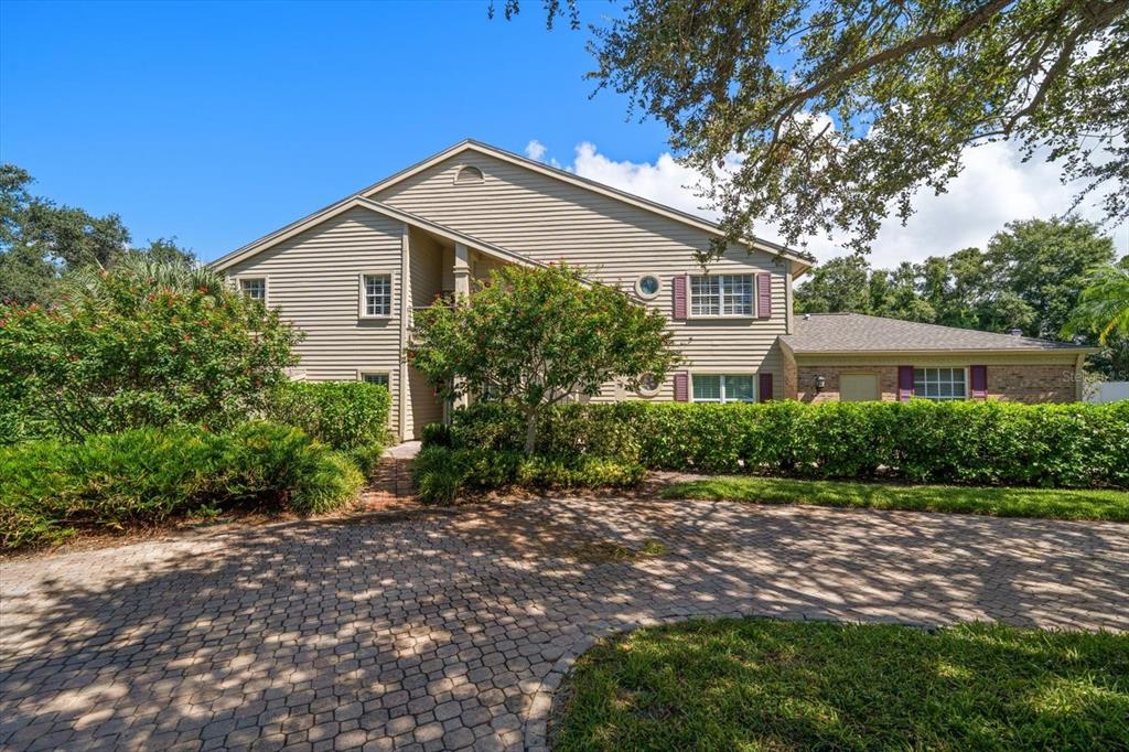 A two-story house with beige siding and a brick-paved driveway surrounded by lush landscaping — 75 PELICAN PLACE, BELLEAIR, FL
